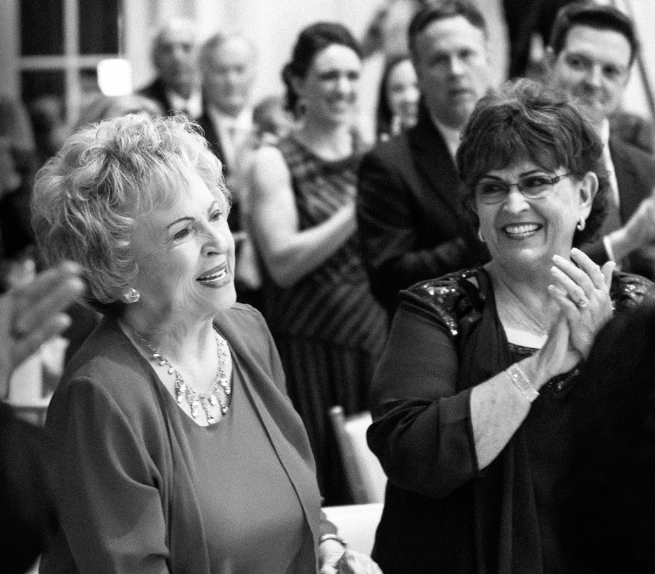 Black white photo of a room full of white men and women-presenting people applauding as they look over at an older-presenting woman, standing at the front of the room