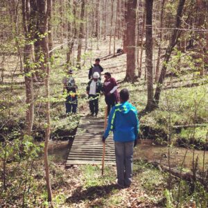Group of five African American people, a mix of elders and younger-presenting people, crossing a bridge over a creek on a forested trail. Two people are holding walking sticks and a younger woman-presenting person is helping an older woman-presenting person walk across the bridge by holding her hand