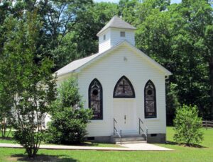 Small white church building with a short single steeple and marbled purple and white stained glass in the front three windows and a cross in the center window panel. The lawn is mowed short with bushes in front, surrounded by green trees during the warmer months