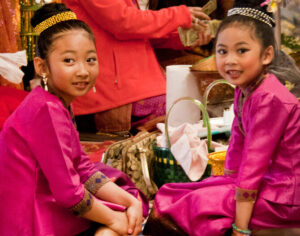 Two young Asian American girls are seated on the ground wearing elegant fuchsia shirts and bottoms that have a satin sheen. Each girl's hair is in a bun on top of their head, surrounded by a metallic band.