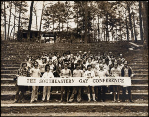 A black and white photograph of a group of young people presenting as majority white and mixed gender, though majority masculine-or man-presenting, posing together outdoors behind a large white banner with dark lettering, reading: "The Southeastern Gay Conference" 