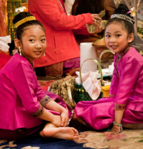 Two young Asian American girls are seated on the ground wearing elegant fuchsia shirts and bottoms that have a satin sheen. Each girl's hair is in a bun on top of their head, surrounded by a metallic band.