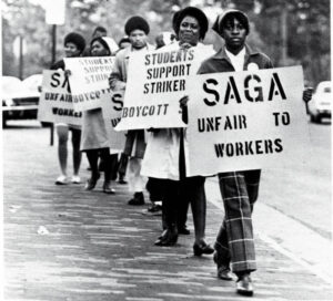 Black and white photo of a picket line of a group of Black majority women-presenting people holding protest signs reading "SAGA - unfair to workers" and "Students Support Strikers - Boycott __ [unreadable]"