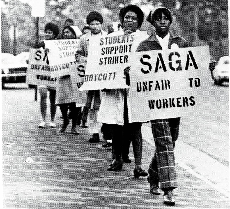 Black and white photo of a picket line of a group of Black majority women-presenting people holding protest signs reading "SAGA - unfair to workers" and "Students Support Strikers - Boycott __ [unreadable]"