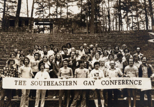 A black and white photograph of a group of young people presenting as majority white and mixed gender, though majority masculine-or man-presenting, posing together outdoors behind a large white banner with dark lettering, reading: "The Southeastern Gay Conference"