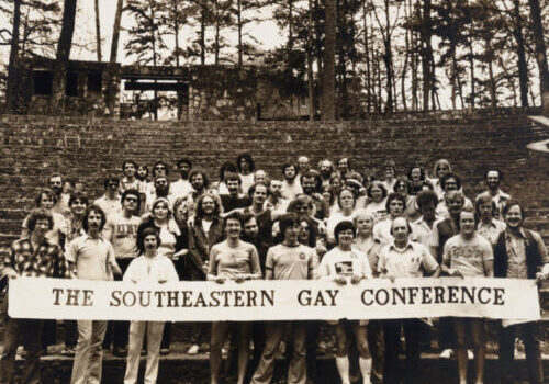 SEGC(1) A black and white photograph of a group of young people presenting as majority white and mixed gender, though majority masculine-or man-presenting, posing together outdoors behind a large white banner with dark lettering, reading: "The Southeastern Gay Conference"